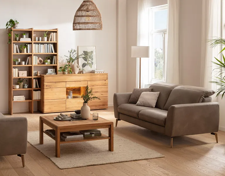 Gemütliches Wohnzimmer im Naturstil mit grauem Ledersofa, Bücherregal und Sideboard aus massiver Eiche, Rattanlampe, vielen Pflanzen und warmem Tageslicht.